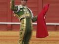 Matador in traditional green and gold bullfighting costume, raising his hand in a gesture of respect or farewell, with a crowd of spectators in the background.