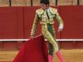 Un torero en traje tradicional de color verde y dorado, con una saya roja y guantes rosa, realiza un gesto en la plaza de toros. La audiencia observa desde los palcos detrás del muro rojo.