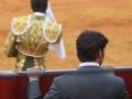 Un torero en traje tradicional de color dorado y blanco, con un pañuelo blanco en la espalda, se prepara para el toro. Detrás de él, un hombre en traje negro sostiene una barra roja. El fondo es una arena de toros con suelo amarillo.