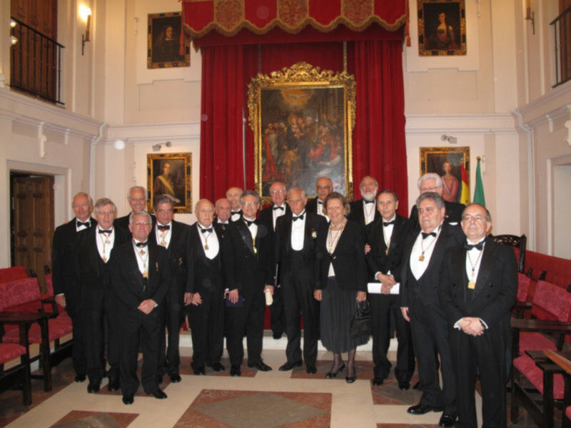 Un grupo de hombres vestidos con trajes formales posan en una sala llena de retratos. La decoración incluye un gran cuadro dorado y cortinas rojas.