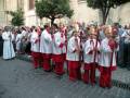 Procesión con niños vestidos de sacerdotes portando candelabros en una calle adoquinada, rodeados por espectadores.
