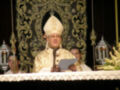 Un sacerdote vestido con un hábito blanco y una mitra dorada, sostiene un libro y se encuentra en frente de un altar con flores y velas. La imagen muestra una ceremonia religiosa, posiblemente un servicio o misa.