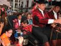 Una imagen festiva de una plaza con toreros, espectadores y un caballo. El personaje central lleva trajes tradicionales rojos con sombrero de copa y guantes. La gente observa atentamente, algunas tomando fotos con sus teléfonos.