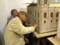 Un hombre en una silla de pieza de madera mira a otro hombre sentado que está trabajando en una maqueta de una torre medieval. La maqueta tiene detalles como ventanas y una aguja con un tejido de lana. La escena ocurre en un entorno interior, posiblemente una oficina o estudio de modelismo.