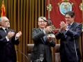 Tres hombres en trajes celebran un premio con aplausos y una estatuilla en una ceremonia formal.