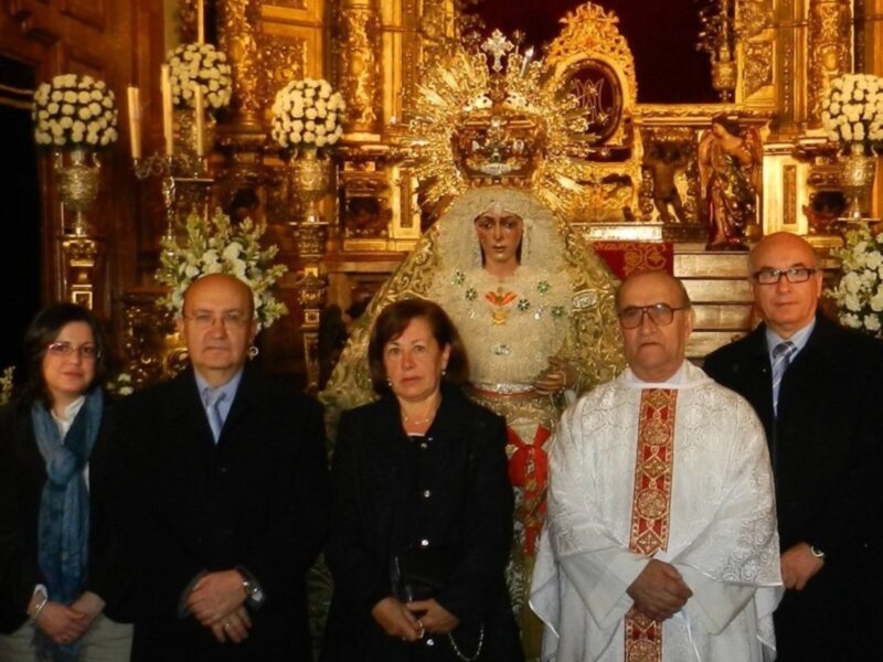 Una imagen festiva donde una persona vestida de blanco y con un cíngulo rojo se encuentra junto a otros cuatro individuos, todos vestidos formalmente. En el fondo, una figura religiosa con un manto dorado y flores blancas se destaca entre adornos dorados. La escena parece ser parte de una celebración religiosa o procesión.