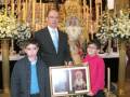Fotografía de una familia en un ambiente religioso, posando junto a un altar con adornos y una imagen de la Virgen María. El padre sostiene un marco con una foto, mientras los hijos miran hacia la cámara. La escena transmite un sentimiento de familia y devoción religiosa.