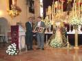 En una iglesia, dos hombres están entregando un reconocimiento a una estatua de la Virgen María. La imagen muestra detalles religiosos y un altar con flores blancas, creando una atmósfera de respeto y honor.