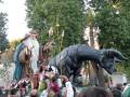 Una figura vestida con trajes tradicionales y tocado se encuentra en el centro de una carroza, rodeada por otros personajes. A su lado, hay un enorme dragón de escultura en piedra. La escena se desarrolla en un ambiente festivo, con árboles y cielo despejado al fondo.