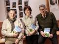 Tres personas sonrientes sostienen libros en una presentación de Casa del Libro. El ambiente es amistoso y el fondo muestra carteles con información sobre la casa del libro.