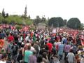 Una multitud de personas con banderas rojas y blancas se congrega en una plaza, frente a un edificio histórico con torres altas. La gente está apretada y parece estar en una manifestación o celebración importante.