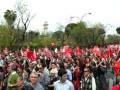 Una multitud con pancartas rojas y banderas ondeando en un parque, con árboles y una torre de control al fondo.