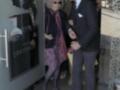 Una mujer anciana con vestido largo y corbata de seda, caminando junto a un hombre en traje negro, ambos miran hacia la izquierda. Detrás de ellos hay una puerta con un cartel que dice "Ateneas".
