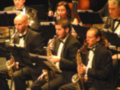 Un grupo de músicos en un concierto, tocan instrumentos como saxofones y clarinetes. El director de orquesta está al frente, gestando con las manos. La escena transmite la energía y concentración de un ensayo o concierto musical en curso.