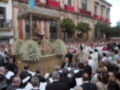 Una procesión religiosa con una carroza decorada con flores blancas, acompañada de fieles y sacerdotes en trajes formales.