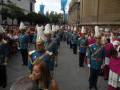 Una procesión militar con soldados en uniformes azules y dorados, caminando por una calle adoquinada. El ambiente es festivo con banderas azules y turquesas en el fondo, y espectadores observando la ceremonia.