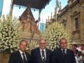 Tres hombres en trajes formales posan frente a un carroza decorada con flores blancas, en una procesión religiosa.