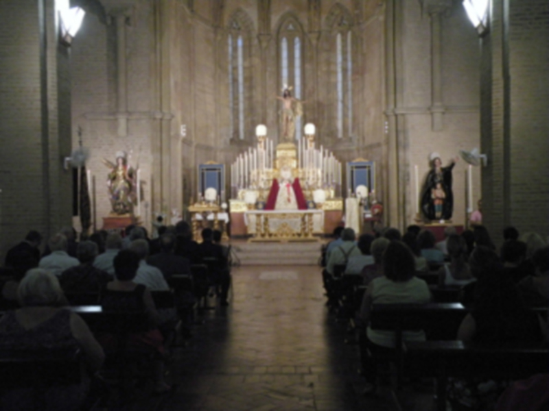 Interior de una iglesia con altares, estatuas y una gran imagen central. La audiencia está sentada en filas de sillas, observando la escena religiosa. La iluminación es suave y cálida, destacando los detalles de la arquitectura y decoración.