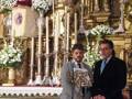 Dos hombres en trajes formales, uno con corbata azul y otro con una camisa blanca y corbata roja, están frente a un altar decorado con flores y una imagen religiosa en el fondo.