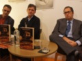 Tres hombres sentados en una mesa con libros y una botella de agua, posiblemente en una conferencia o presentación de libros.