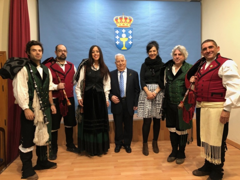 Una imagen de una reunión formal con siete personas vestidas en trajes tradicionales. La pared detrás muestra un escudo de armas con una corona y estrellas, probablemente representando la bandera de Galicia.