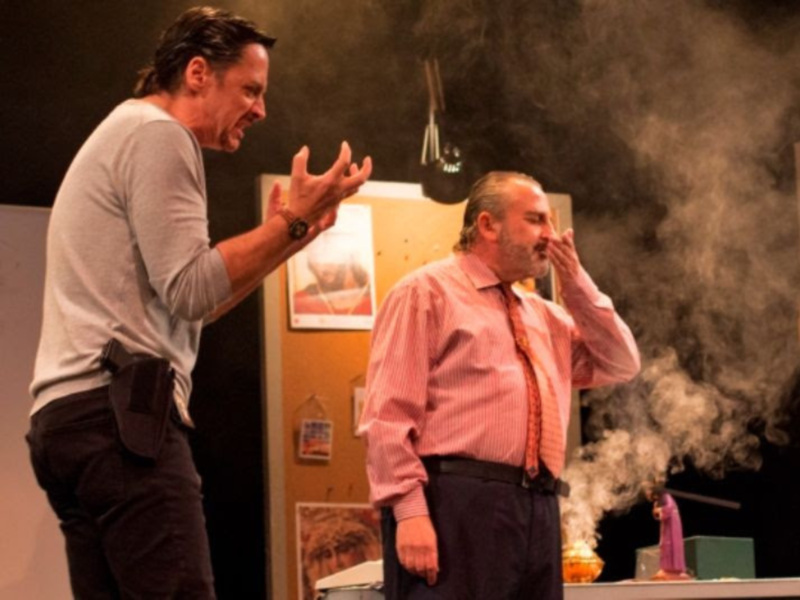 Dos hombres en una escena teatral, uno con una camisa blanca y el otro con una camisa rosa y corbata, con humo en el fondo.