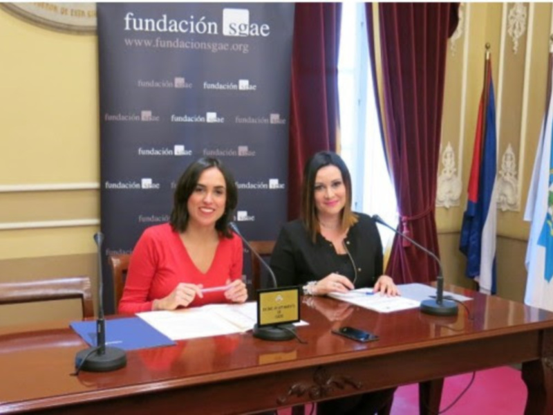 Dos mujeres firman un documento en una sala de conferencias, con banderas y la web de la Fundación SGAEE visible en el fondo.