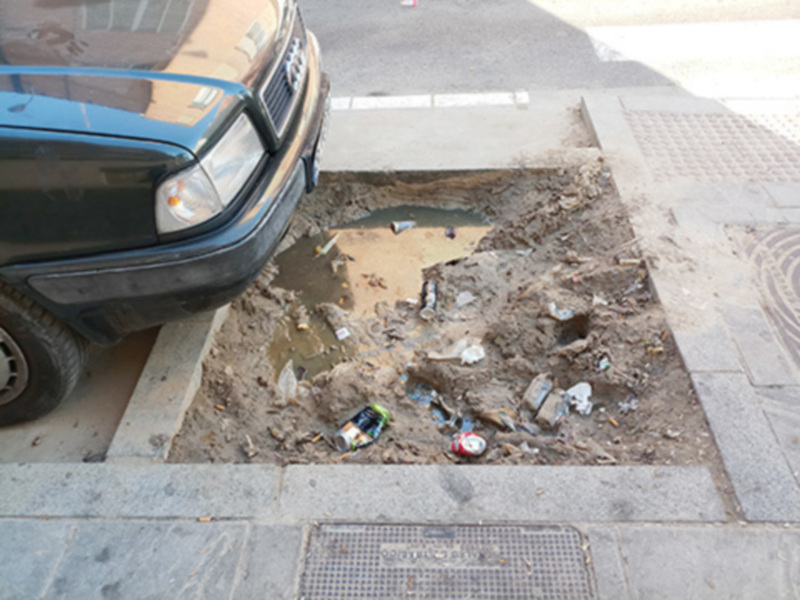 Se observa un coche estacionado en una calle con desperdicios y basura acumulados en el suelo, cerca de un agujero en la calzada. La escena sugiere problemas de limpieza y mantenimiento en el área.