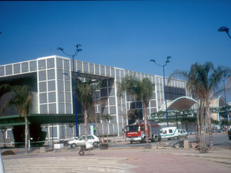 Edificio moderno con estructura de vidrio y metal, ubicado en una zona con árboles y vehículos a la entrada. El cielo es azul claro y el entorno parece ser un lugar urbano o industrial.