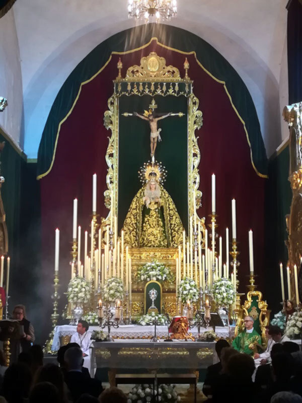 Imagen de una iglesia con un altar dorado y una imagen central. En el fondo, hay una cruz con un crucificado y una figura dorada en la parte superior. La imagen está iluminada por las velas encendidas y una lámpara de araña. La decoración es elegante con detalles dorados y un fondo rojo.