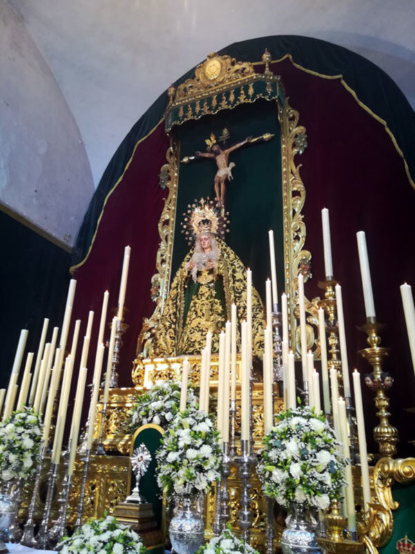 Altar con imagen de la Virgen y Cristo crucificado, adornado con flores y candelabros, en un entorno religioso.