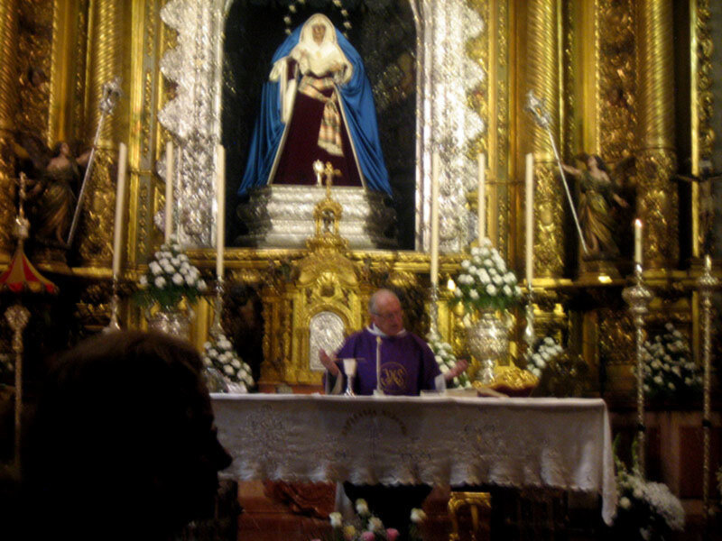 Un altar de una iglesia con una estatua de la Virgen María en el centro, rodeada por flores y velas. En el fondo, se puede ver una figura humana vestida con un traje religioso y sosteniendo una cruz. El altar está adornado con detalles dorados y cristales, creando un ambiente de reverencia.
