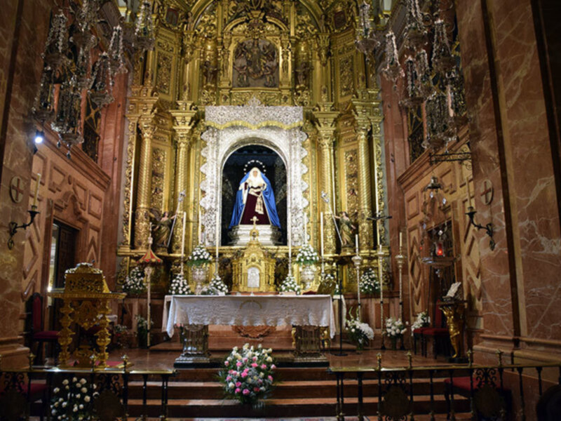 Interior de una iglesia con altares dorados, flores en macetas y un altar central.