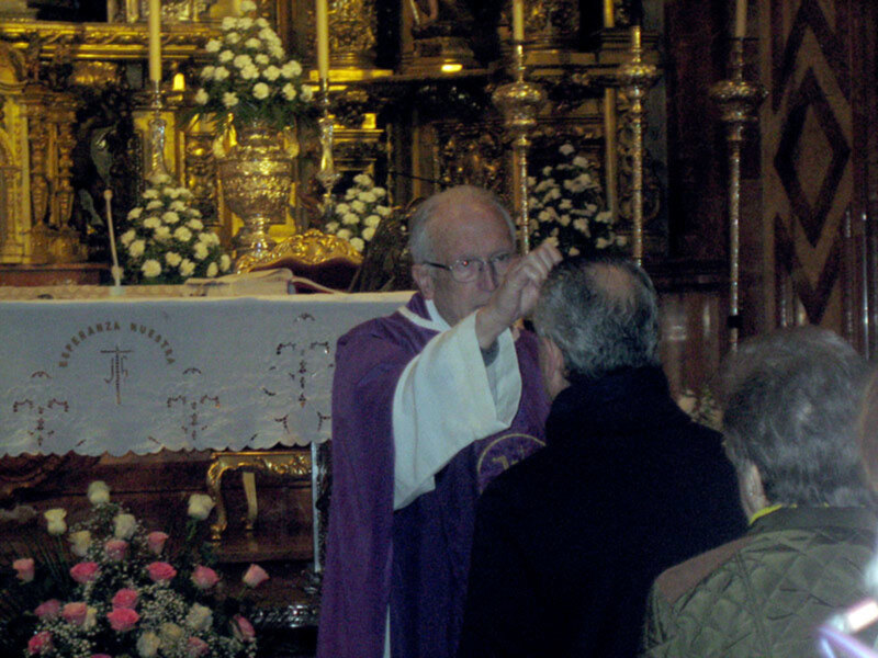 Imagen de una ceremonia religiosa en un altar dorado con flores blancas y rosadas. Un hombre vestido de rojo celebra la ceremonia, mientras que una persona en negro observa. La imagen transmite un ambiente de respeto y solemnidad.
