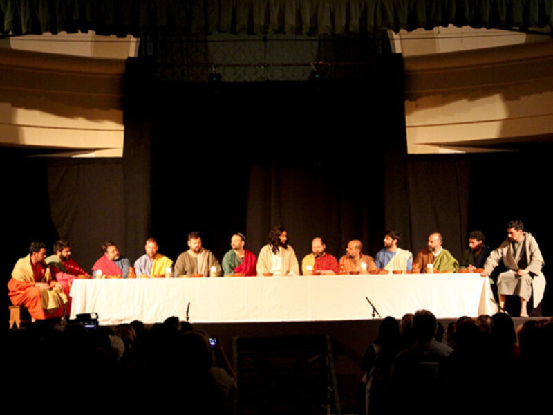 Una escena de teatro con un grupo de actores sentados en una mesa, vestidos con trajes coloridos. La iluminación es suave y la atmósfera parece ser de una representación teatral.
