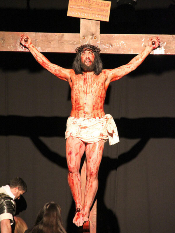 Un Cristo crucificado en una cruz de madera, con detalles rojos y ojos abiertos. En la parte superior de la cruz hay un cartel con texto en inglés que dice "Jesus Christ". La imagen se toma en una escena con un fondo oscuro y algunas personas a la izquierda.