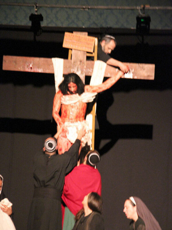 Una escena teatral dramática muestra a un hombre en una cruz, con otros personajes vestidos de negro alrededor. La escena parece ser parte de una representación teatral religiosa o histórica.