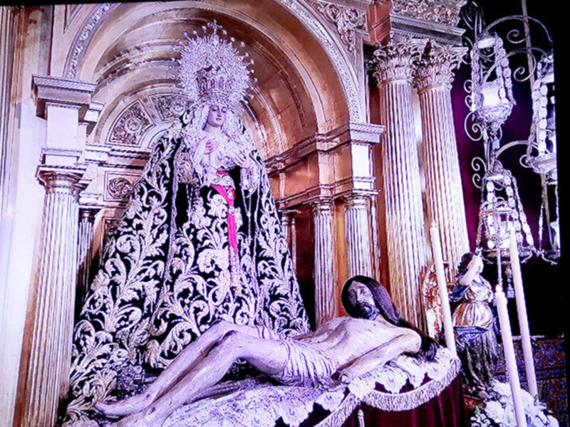 Imagen de una estatua religiosa en un interior de iglesia, con un personaje en posición de oración y una figura más baja a su lado. La escena está iluminada por la luz natural que entra desde un ventanal no visible en la imagen. La estatua está rodeada por detalles arquitectónicos y decorativos, como columnas y molduras.