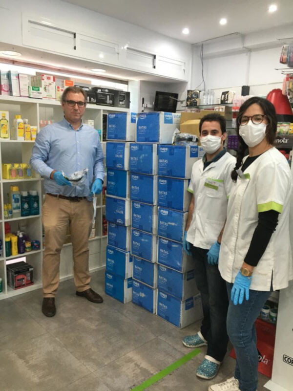 Dos personas posan frente a una estantería llena de cajas azules en un entorno de laboratorio o almacén. La persona a la izquierda lleva una camisa azul claro y pantalones beige, mientras que la persona a la derecha está vestida con una camiseta blanca y pantalones negros. Ambos tienen guantes en las manos, lo que sugiere un ambiente de trabajo con medidas de seguridad. La estantería contiene diversos productos, incluyendo botellas y cajas de diferentes tamaños.