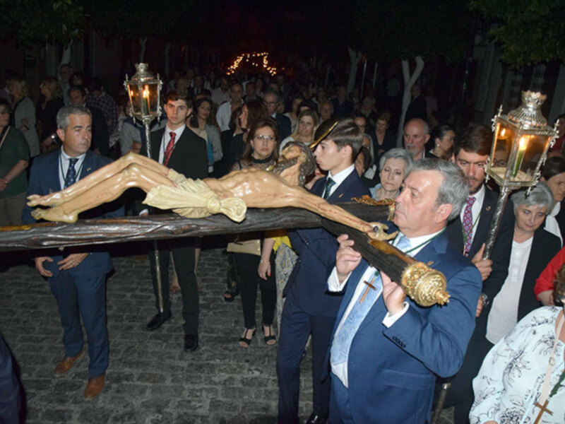 Una procesión religiosa con una imagen de Cristo crucificado siendo llevada por varios hombres en trajes formales. La gente observa con atención, algunos sostienen faroles. El ambiente es solemnemente festivo y tradicional.