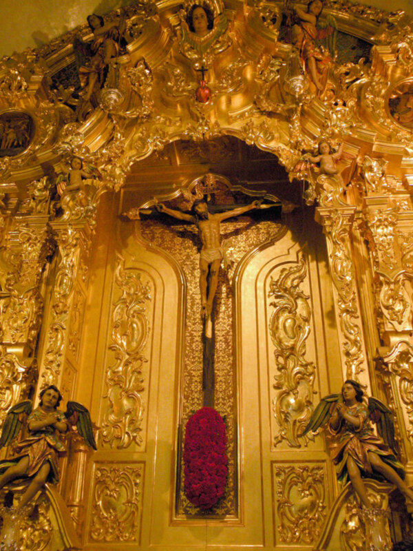 Imagen de una iglesia con un altar dorado elaborado, en el centro hay una cruz con un Cristo crucificado. A ambos lados del altar se encuentran figuras de ángeles con alas, una figura masculina y otra femenina. La imagen está iluminada por la luz que proviene de arriba, creando sombras y destellos en los detalles dorados.