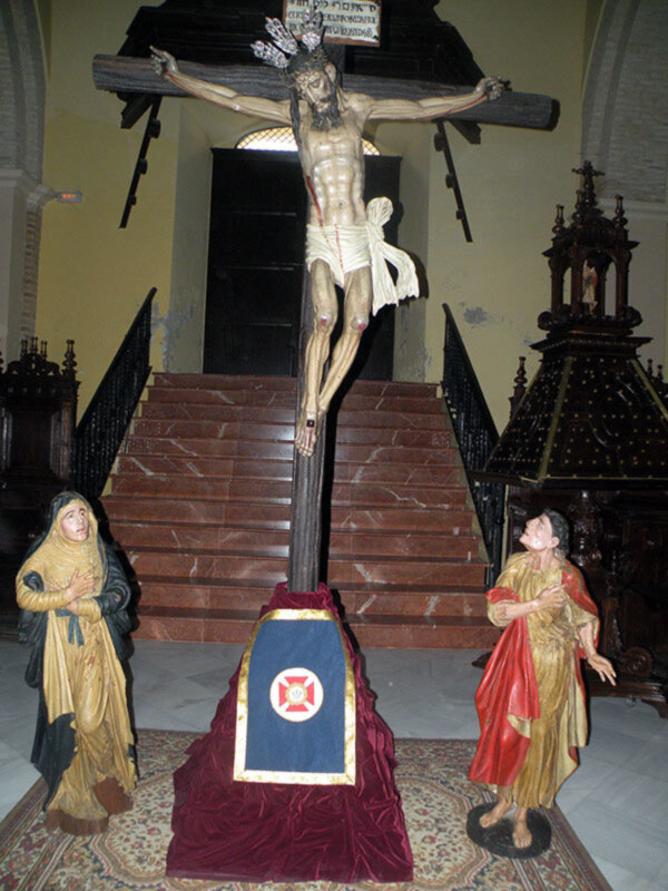 Estatua de Jesús crucificado en una iglesia, con una figura de mujer en la base y una figura de mujer en el lado.