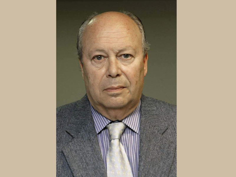 Un hombre de cabello blanco y barba, con una expresión seria. Lleva un traje gris claro, camisa de rayas y corbata blanca. El fondo es un tono claro, probablemente beige o marrón claro.