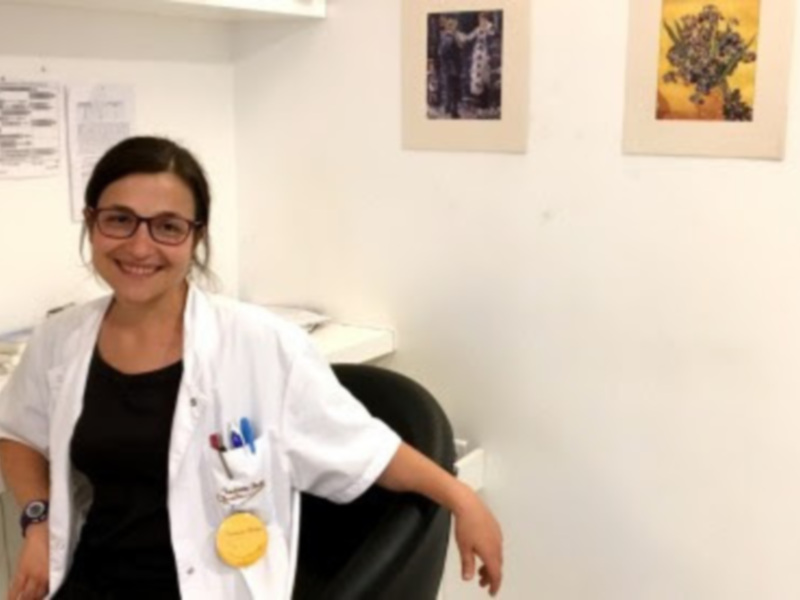 A una persona en un entorno de oficina o consulta médica, con documentos y cuadros decorativos en el fondo. La persona está vestida con un uniforme médico, incluyendo una chaqueta blanca y un distintivo amarillo en el pecho. La persona sonríe, lo que sugiere una atmósfera positiva y acogedora.