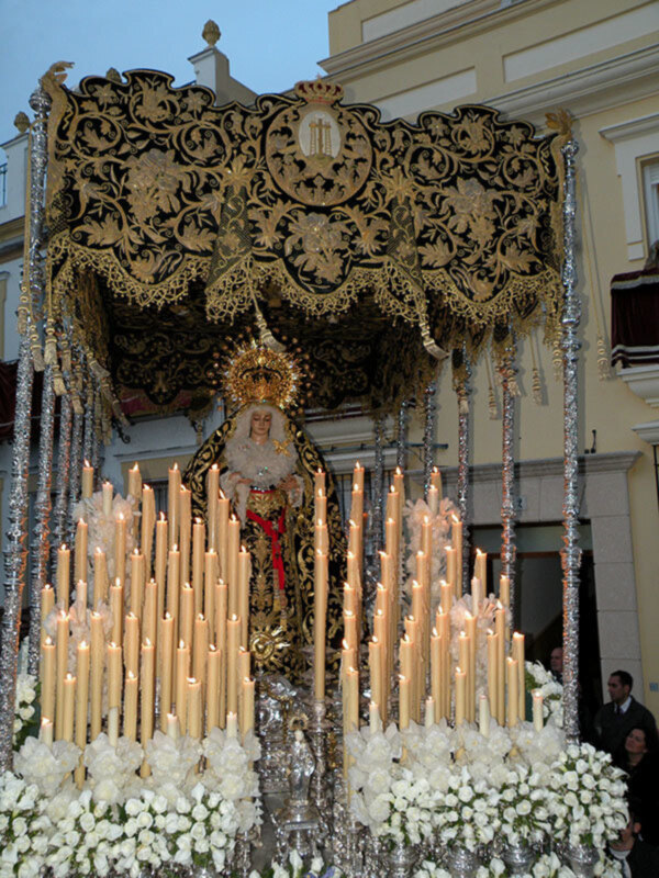 Una imagen de una procesión religiosa con un altar adornado, velas encendidas y flores blancas. La imagen muestra una escena festiva o ceremonial, probablemente relacionada con una celebración religiosa.