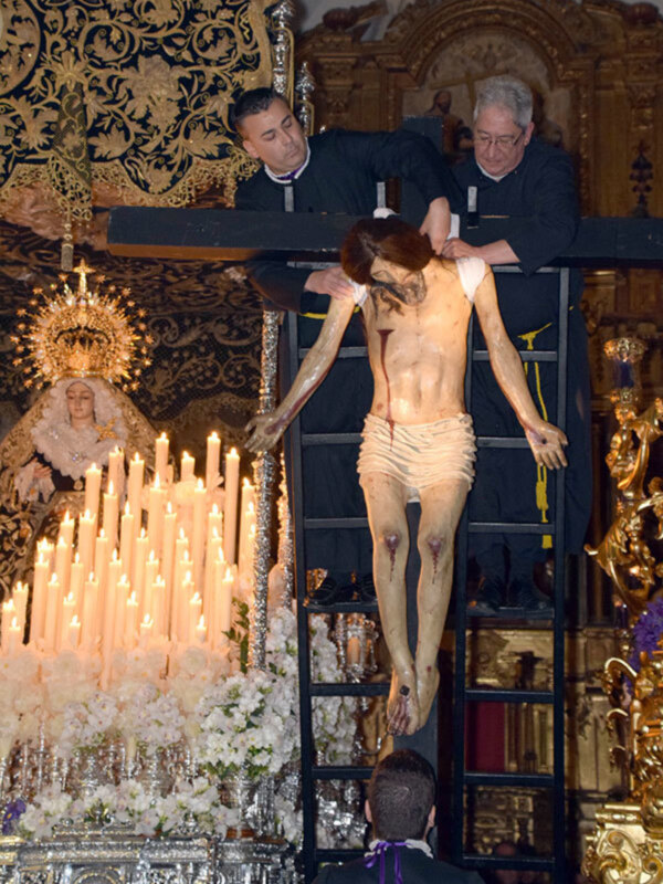 Imagen de una procesión religiosa con un crucificado en el centro, rodeado por velas encendidas y flores. En el fondo se observa un altar con una imagen de la Virgen María. La escena transmite solemnidad y devoción cristiana.