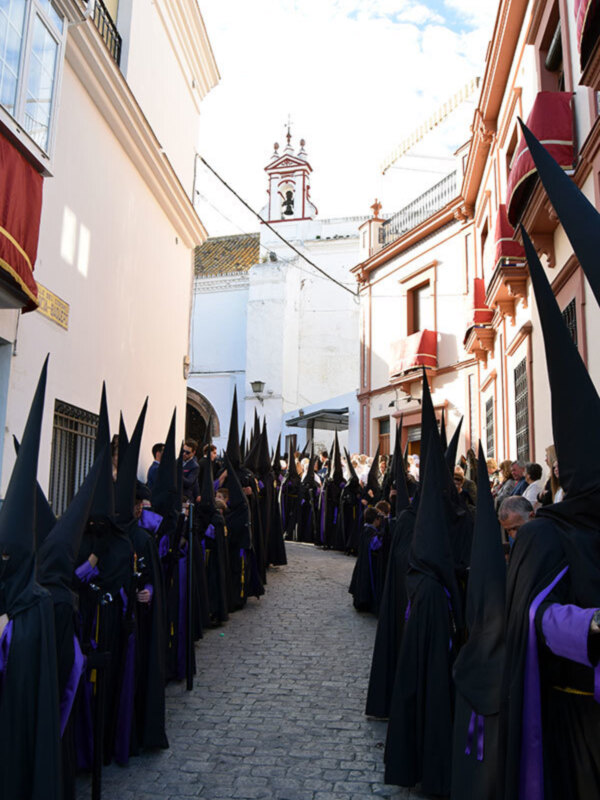 Procesión de penitentes en una calle de la ciudad, con capas negras y capuchones, frente a edificios históricos con balcones rojos y azules.