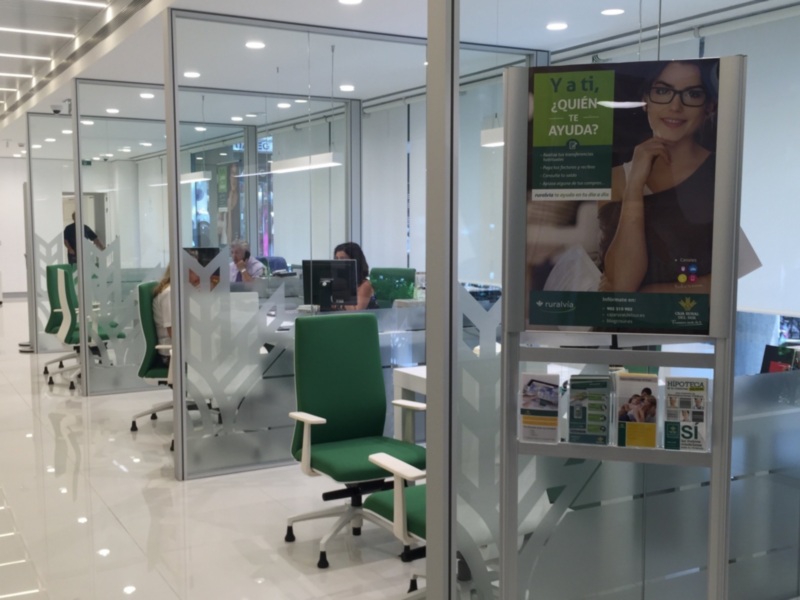 Interior de una oficina moderna con mesas de trabajo, sillas verdes y paneles divisorios de vidrio. En la pared, un cartel promocional de una entidad financiera.