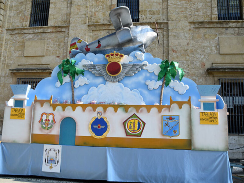 Escultura temática con avión, palmeras y escudos militares, frente a un edificio histórico.