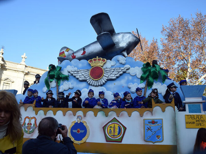 El pié de foto muestra una imagen festiva con un carro de carnaval decorado como un barco militar. El carro está pintado en azul y blanco, con detalles de palmeras verdes y un escudo real. En la parte superior del carro, hay una figura de un barco con una torre y una bandera. Debajo del carro, se pueden ver varios personajes vestidos con uniformes militares y sombreros. El cielo es azul claro, y en el fondo se pueden ver árboles con hojas amarillas.